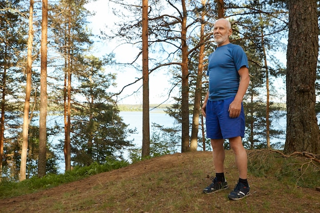 Activo guapo maduro sizty año viejo vistiendo elegante ropa deportiva de pie sobre la hierba seca en el bosque contra los árboles y el vasto lago. concepto de energía, naturaleza, envejecimiento y jubilación