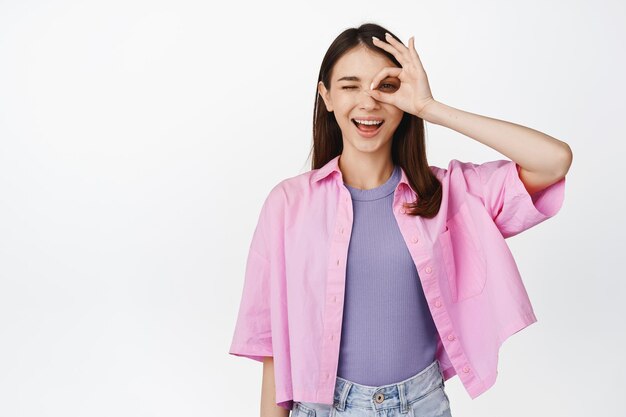 De acuerdo, la chica morena sonriente muestra el signo de ok y guiña un ojo sin ningún problema, gesto cero de pie sobre fondo blanco