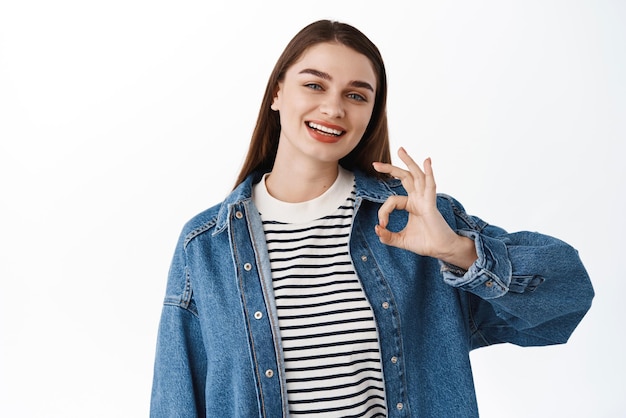 De acuerdo, está bien La niña feliz sonriente muestra la aprobación de la señal OK, da una respuesta positiva, dice que sí, asiente y recomienda la calidad de la garantía del evento de pie sobre un fondo blanco