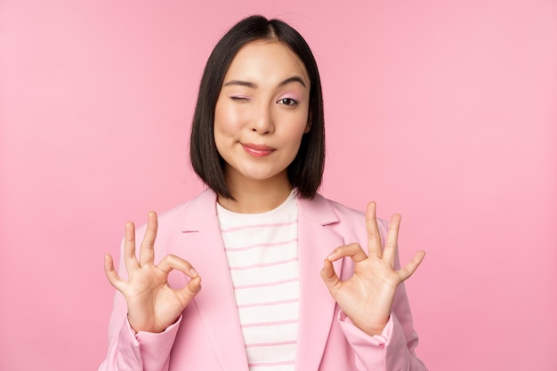 De acuerdo, excelente mujer de negocios en traje corporativo que muestra un gesto de aprobación aceptable recomendando algo dar retroalimentación positiva y sonriendo complacido posando sobre fondo rosa