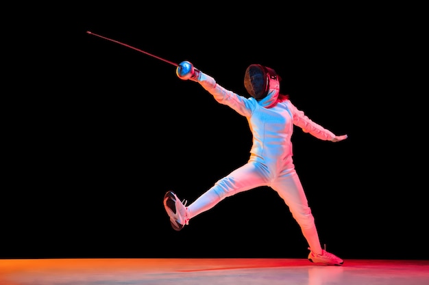 Hacia adelante. Jovencita en traje de esgrima con espada en mano aislada sobre fondo negro, luz de neón. Joven modelo practicando y entrenando en movimiento, acción. Copyspace. Deporte, juventud, estilo de vida saludable.