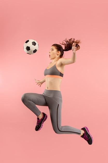 Adelante a la victoria. La joven como futbolista saltando y pateando la pelota en el estudio sobre un fondo rojo. Aficionado al fútbol y concepto de campeonato mundial. Conceptos de las emociones humanas