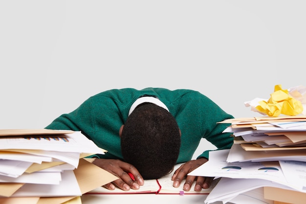 El adicto al trabajo estresante mantiene la cabeza gacha sobre el escritorio, se siente cansado y con exceso de trabajo, tiene mucho trabajo, se prepara para el próximo examen, escribe información en el diario