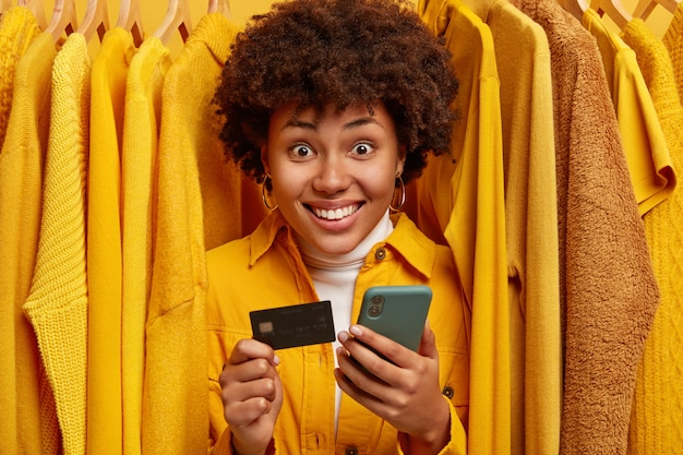Adicto a las compras de raza mixta positiva se encuentra cerca del estante con ropa, usa tarjeta de crédito y teléfono inteligente para realizar compras