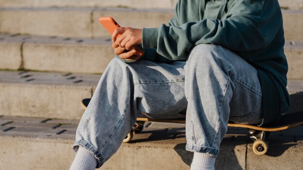 Foto gratuita adolescente afuera sentado en patineta y sosteniendo el teléfono inteligente