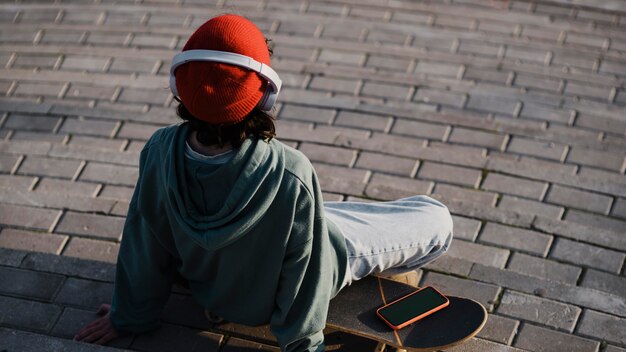 Adolescente al aire libre escuchando música con auriculares mientras está sentado en patineta