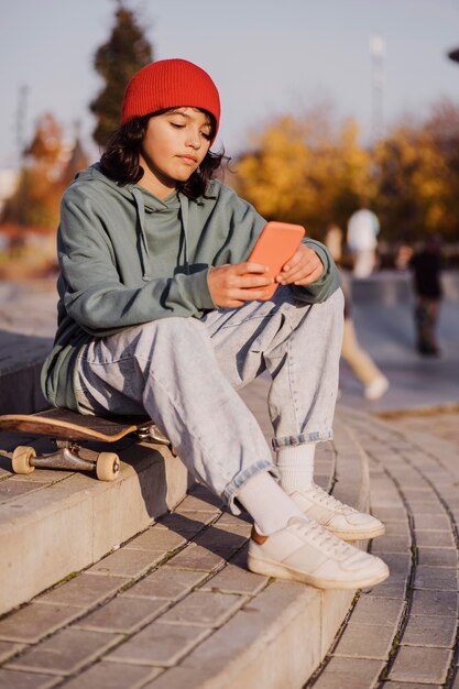 Adolescente al aire libre sentado en patineta y sosteniendo el teléfono inteligente