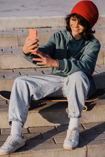 Adolescente al aire libre sentado en patineta y tomando selfie