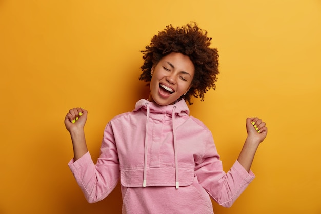 Adolescente alegre baila despreocupado, se siente optimista, es campeón, hace chocar los puños, cierra los ojos y sonríe ampliamente, se viste con una sudadera informal de terciopelo, se regocija por el triunfo, se mueve contra la pared amarilla.