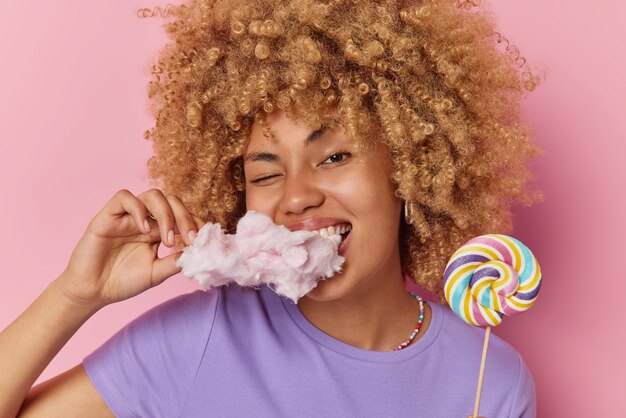 Una adolescente alegre y despreocupada disfruta comiendo dulces, bocados de algodón de azúcar, sostiene una piruleta en un palo, viste una camiseta púrpura casual aislada sobre un fondo rosa Concepto de adicción al azúcar y alimentos poco saludables