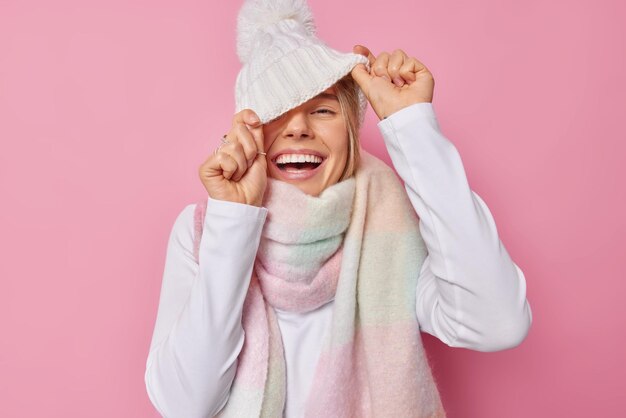 Una adolescente alegre se divierte esconde la cara detrás de un gorro de punto blanco tiene un estado de ánimo optimista, usa un suéter y una bufanda alrededor del cuello, sonríe ampliamente, tiene dientes perfectos, incluso los tontos están llenos de energía