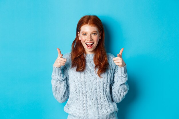 Adolescente alegre mostrando aprobación, hacer pulgares hacia arriba y estar de acuerdo, que le gusta algo, de pie sobre fondo azul.