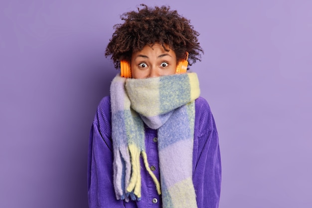 Foto gratuita adolescente alegre con el pelo rizado envuelto en una bufanda pasa su tiempo libre caminando al aire libre durante el día de invierno escucha una agradable melodía.