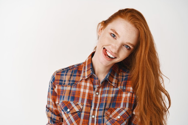Adolescente alegre con pelo rojo y piel pálida mostrando la lengua y sonriendo feliz a la cámara de pie sobre fondo blanco.