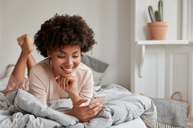 Adolescente alegre de piel oscura con cabello afro, vestida con ropa de dormir, utiliza teléfono móvil