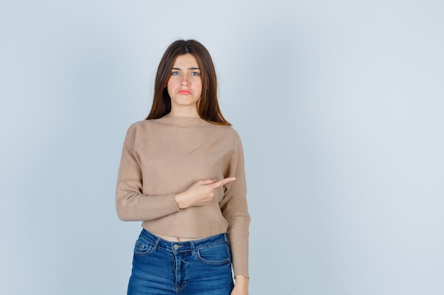 Adolescente apuntando hacia la derecha, curvando el labio inferior en suéter, jeans y mirando triste, vista frontal.