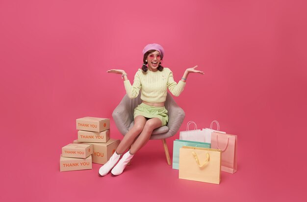 Una adolescente asiática feliz sentada en un sofá con bolsas de compras y una caja de regalo aislada de fondo rosa