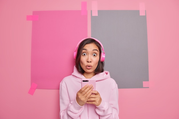 Una adolescente asiática sorprendida usa los auriculares inalámbricos más nuevos usa una aplicación musical móvil escucha una pista de audio escucha noticias sorprendentes vestida casualmente aislada sobre una pared rosa