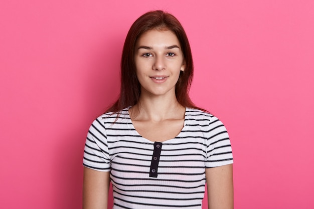 Adolescente atractiva joven con camiseta casual a rayas, posando contra la pared rosa, con una sonrisa encantadora, parece feliz.