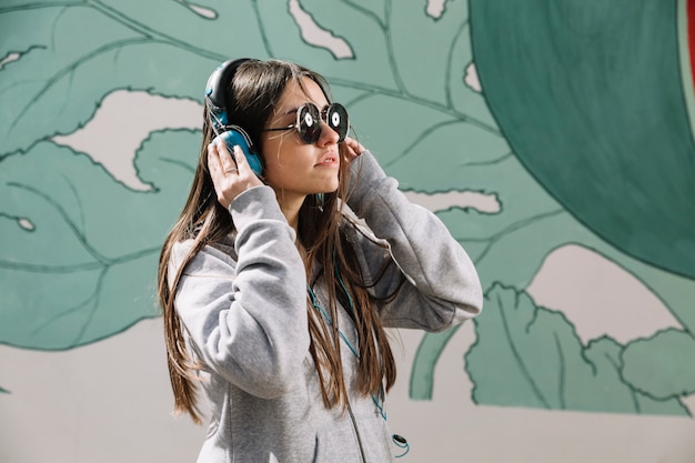 Adolescente con auriculares y gafas de sol delante de la pared pintada