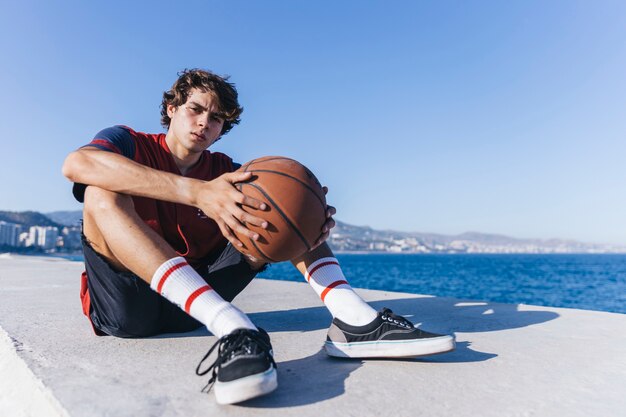 Adolescente con baloncesto sentado cerca del mar