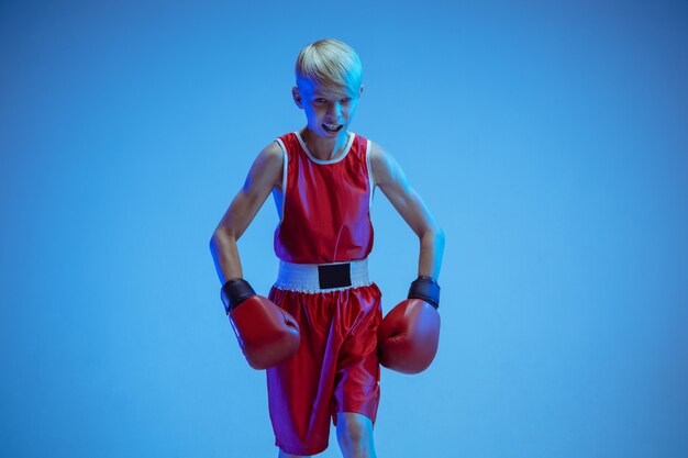 Adolescente en boxeo de ropa deportiva aislado sobre fondo azul de estudio en luz de neón. Boxeador caucásico masculino novato entrenando duro y trabajando, pateando. Deporte, estilo de vida saludable, concepto de movimiento.