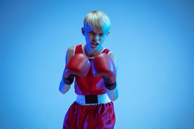 Adolescente en boxeo de ropa deportiva aislado sobre fondo azul de estudio en luz de neón. Boxeador caucásico masculino novato entrenando duro y trabajando, pateando. Deporte, estilo de vida saludable, concepto de movimiento.