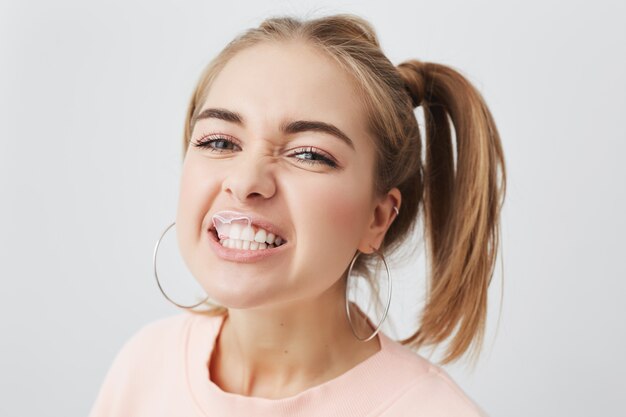 Adolescente burlona, divertida, juguetona, rubia, con dos coletas y grandes aretes redondos, vestida con un suéter rosa que muestra los dientes y el ceño fruncido, aislado