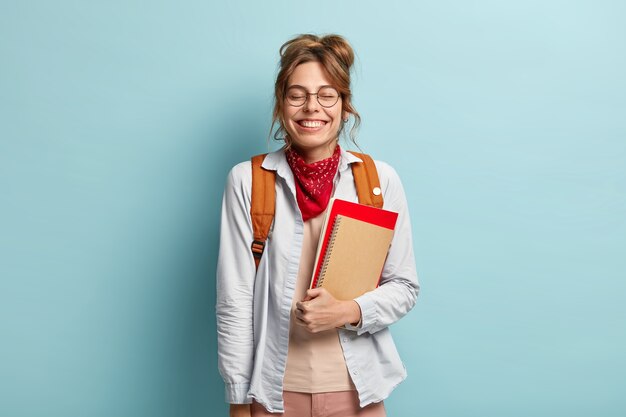 Adolescente complacida se ríe alegremente, mantiene los ojos cerrados, escucha una broma divertida durante el descanso entre clases