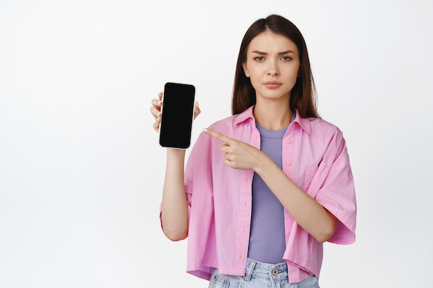 Adolescente decepcionada señalando la pantalla del teléfono inteligente frunciendo el ceño como expresando disgusto emoción disgustada de pie sobre fondo blanco.