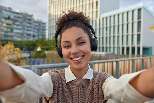 Una adolescente encantadora y positiva con el pelo rizado peinado se toma una selfie en un entorno urbano sonríe alegremente escucha música de playlit usa auriculares inalámbricos en los oídos y está de buen humor durante el día de primavera