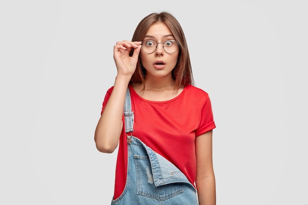 Adolescente con estilo sorprendido en camiseta roja casual y overoles de mezclilla, mantiene la mano en el borde de las gafas