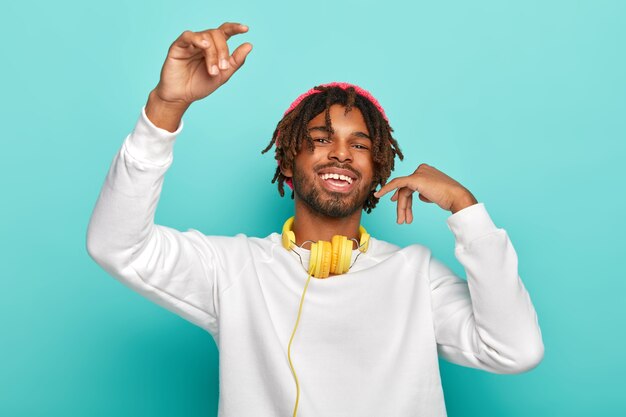 Adolescente feliz encantado con rastas, levanta los brazos, siente alegría mientras escucha su música favorita a través de auriculares, se mueve al ritmo de la canción, usa un jersey blanco