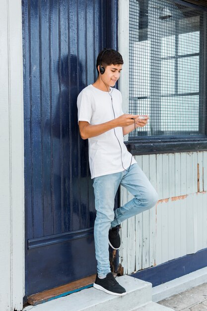 Adolescente feliz que se coloca en la puerta azul cerrada usando el teléfono móvil con el auricular en su cabeza