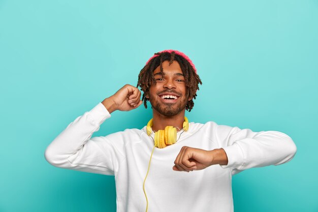 Adolescente feliz tiene rastas cortas, se mueve al ritmo de la música, siente placer, vestido con un suéter blanco, aislado sobre fondo azul