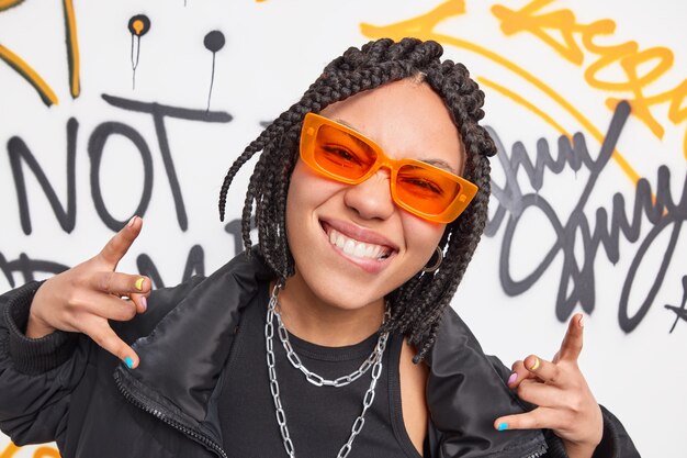 Una adolescente funky de una pandilla callejera hace un gesto genial, muerde el labio inferior tiene trenzas, usa gafas de sol naranjas, una chaqueta negra de moda se divierte en lugares públicos, posa contra una pared de graffiti, tiene un estado de ánimo lúdico