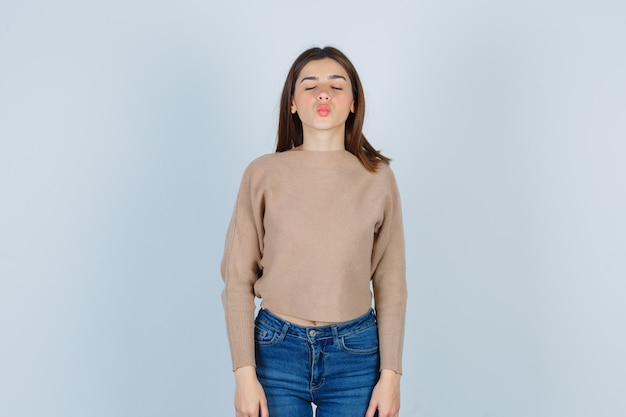 Adolescente haciendo pucheros con los labios, cerrando los ojos en suéter, jeans y mirando tentador, vista frontal.