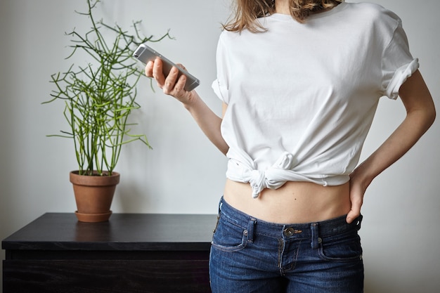 Adolescente irreconocible en camiseta casual y jeans posando en el interior de la acogedora habitación con maceta