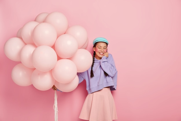 Una adolescente llena de alegría mantiene los ojos cerrados, sonríe ampliamente, muestra los dientes blancos, usa boina, sudadera y falda, sostiene globos inflados, celebra la licenciatura, aislado en la pared rosada