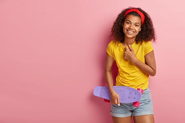 Una adolescente llena de alegría se ríe positivamente, está de buen humor, expresa emociones agradables, se viste con ropa casual de verano, sostiene longboard, se divierte con amigos