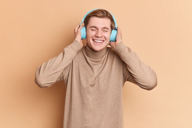 Adolescente masculino alegre relajado con una gran canción usa auriculares estéreo azules en las orejas, tiene una amplia sonrisa y quiere bailar vestido con cuello alto informal