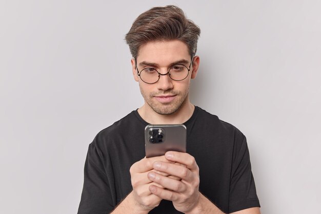 Adolescente masculino guapo serio mira atentamente el teléfono inteligente envía correos electrónicos utiliza la aplicación para chatear cheques notificación usa gafas transparentes camiseta negra aislada sobre fondo blanco