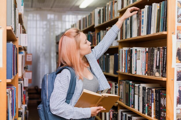 Adolescente con mochila recogiendo el libro de la estantería