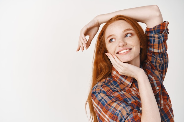 Adolescente mujer soñadora con cabello rojo natural, posando en una pared blanca y mirando hacia la izquierda en el logo, tocando la cara perfecta sin maquillaje