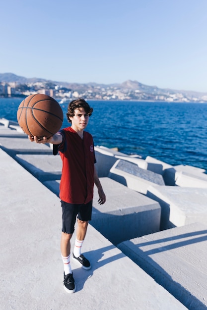 Adolescente de pie cerca del mar mostrando baloncesto