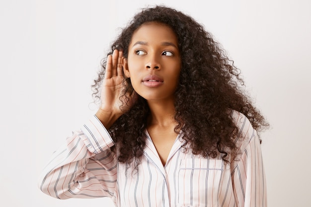 Adolescente de piel oscura en pijama con la mano en la oreja, escuchando a escondidas. Bonita mujer afroamericana con mirada curiosa, escuchando una interesante conversación secreta, manteniendo la boca abierta