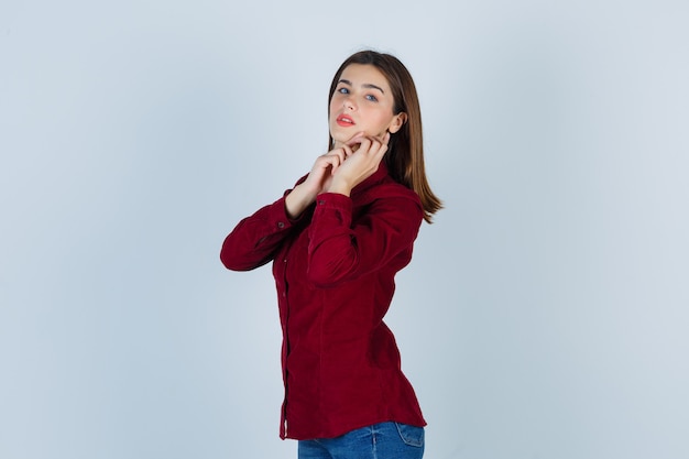 Adolescente posando mientras toca su mandíbula en camisa burdeos y luciendo encantador, vista frontal.