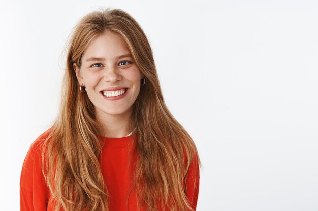 Adolescente positiva con cabello largo natural y pecas sonriendo alegremente con expresión amistosa y extrovertida de pie en una cálida sudadera roja de moda