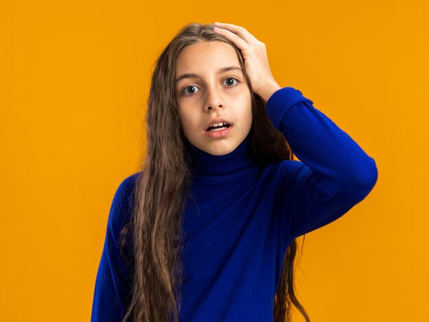 Adolescente preocupada mirando al frente manteniendo la mano en la cabeza aislada en la pared naranja