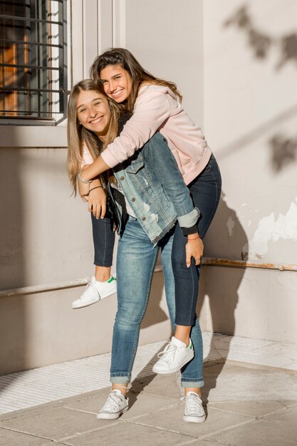 Adolescente que da a su amiga a cuestas paseo de pie delante de la pared